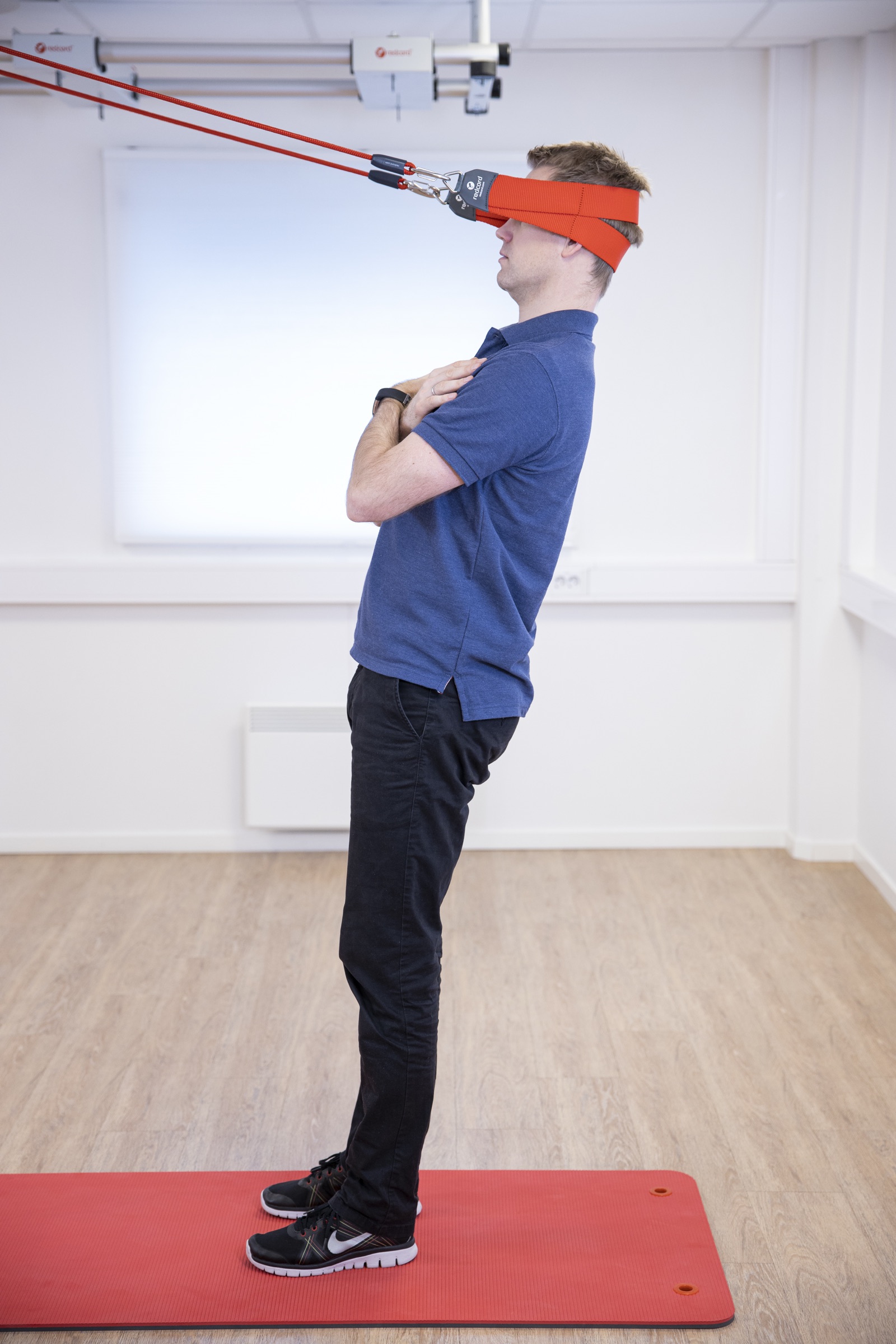 A person performing a supported standing posture-correction exercise with a Redcord head sling